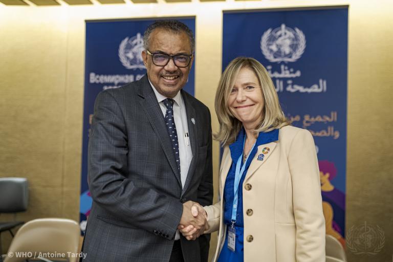 Two individuals, a man and a woman, are shaking hands and smiling in front of WHO banners. The man is in a dark suit and tie, and the woman is in a beige blazer with a blue shirt.