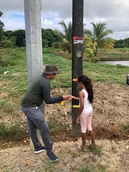 A man wearing a hat and a girl in pink pants measure water levels on a wooden pole marked with colored lines, outdoors near a field with palm trees in the background.