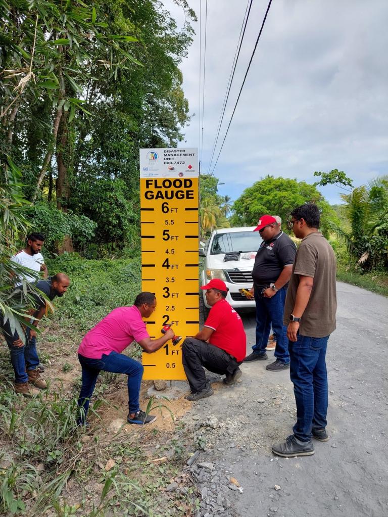 Several people are standing on a roadside next to a newly installed yellow flood gauge sign. One person is adjusting the base of the sign while others observe. A white vehicle is parked in the background.