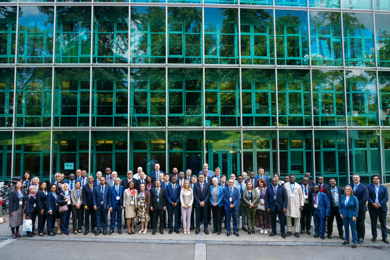 A large group of individuals in formal attire stands in several rows in front of a modern glass building, posing for a group photo.