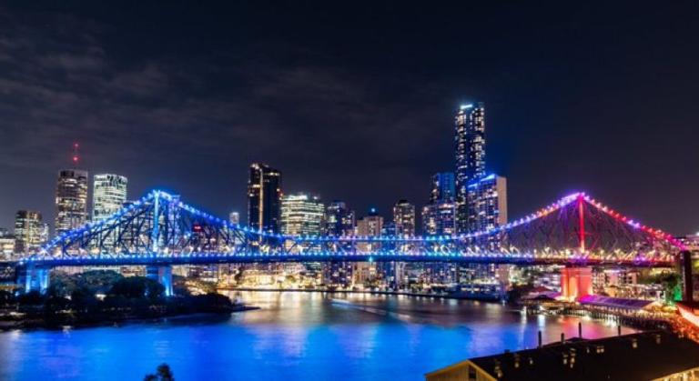 Night view of a brightly lit suspension bridge with blue and red lights, spanning across a river, with a city skyline featuring illuminated high-rise buildings in the background.