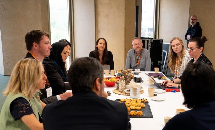 A group of people gather around a table with snacks and laptops, engaging in discussion in a well-lit conference room.