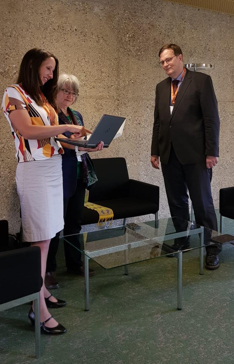 Three people are gathered around an open laptop. One woman is holding the laptop while showing something to the other two individuals. They are standing in a room with a glass table and chairs.