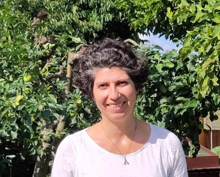 A woman with short curly hair stands smiling in front of lush green foliage on a sunny day, wearing a white top and a necklace with an Eiffel Tower pendant.