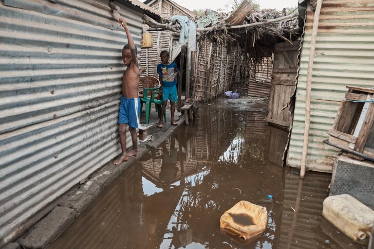 Two boys stand in a flooded alley between metal and wooden structures. One boy is holding onto the wall, and the ground is covered with water and scattered debris, including a yellow container.