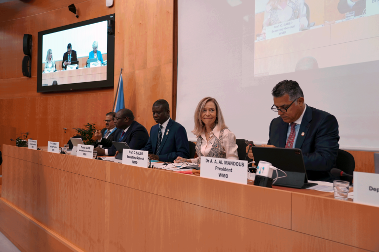 A panel of five people is seated at a long table with nameplates, microphones, and documents in front of them during a conference. A screen above displays a live feed of the session.