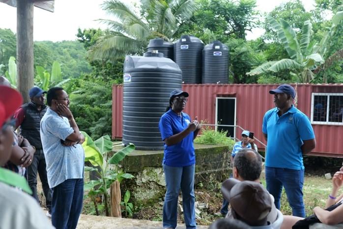 A group of people is gathered outdoors near water storage tanks. Two individuals are speaking, with surrounding greenery in the background.