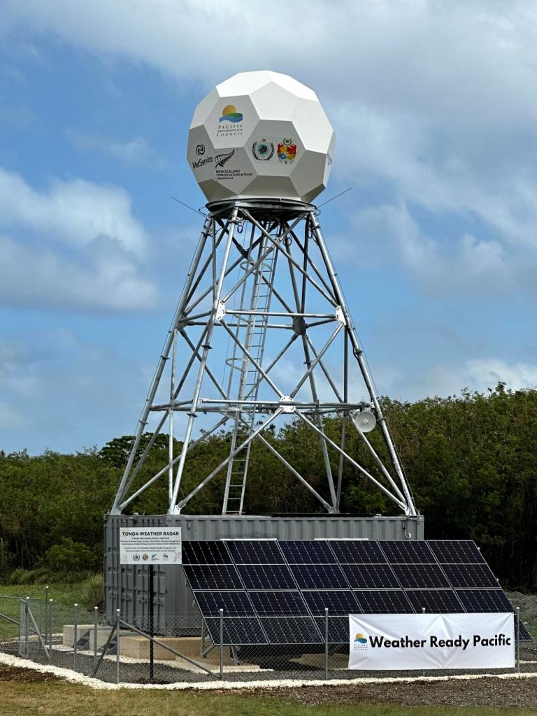 A weather radar station with a geodesic dome atop a metal tower, solar panels at the base, and a banner reading "Weather Ready Pacific." Trees and a cloudy sky are in the background.