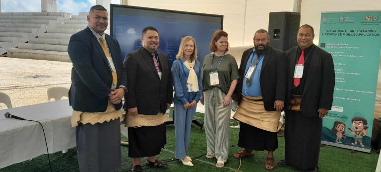 A group of six people stands in front of a display screen at an indoor event. They are dressed in formal attire and nametags. A banner about Tonga's early warning and response mobile application is on the right.