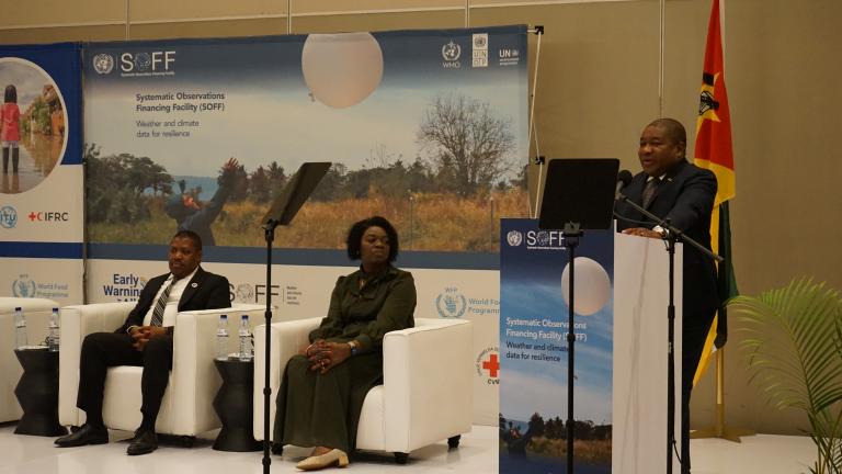 A man speaks at a podium during an event, with two seated individuals and a backdrop displaying text and imagery related to systematic observations and climate. Flags and logos are present in the background.