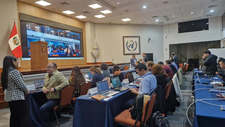 A conference room with people working on laptops, a large screen displays a video conference, and a Peruvian flag is visible. Several attendees are conversing or setting up equipment.