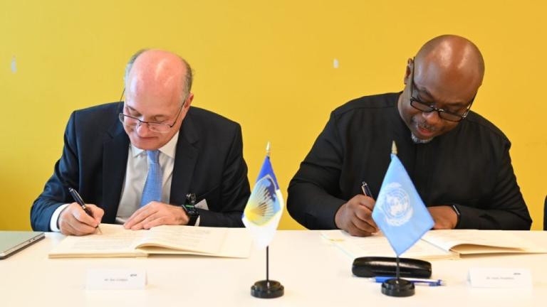 Two men seated side-by-side at a table, both signing documents. In front of them are small table flags representing different organizations, one with a blue flag. The background is a yellow wall.