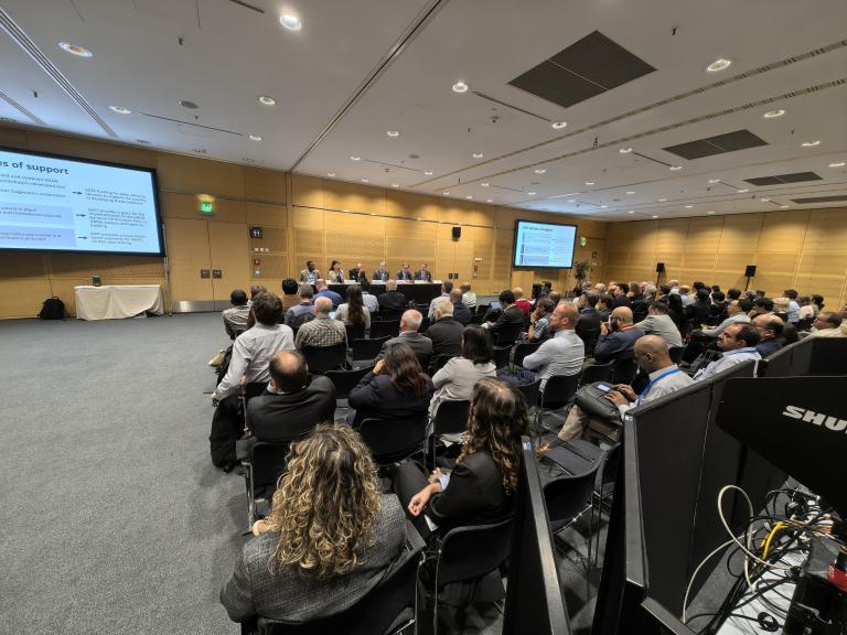 A conference room filled with attendees seated and facing a panel of speakers addressing them from a long table at the front. Presentation slides are projected on screens behind the speakers.