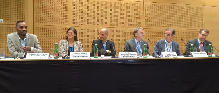 A panel of six professionals sitting at a long table, each with a microphone and water bottle, participating in a conference discussion.