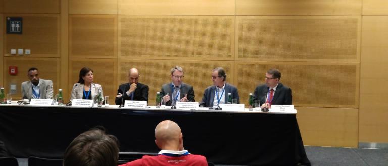 A panel of six people is seated at a long table with microphones and nameplates, participating in a discussion at a conference. A few audience members are visible in the foreground.