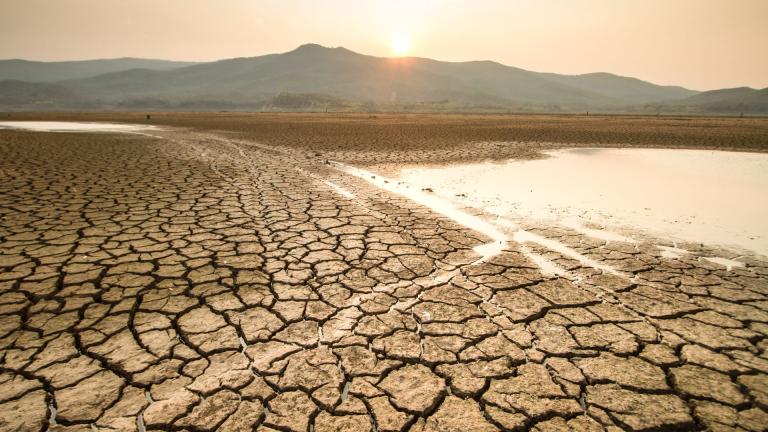 A dry, cracked landscape with a small pool of water in the foreground, and mountains silhouetted against a hazy sunset in the background.