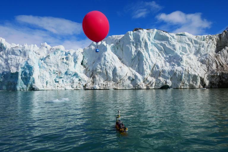 A glacier with a floating red balloon above it and a small, partially submerged device in the water below, set against a blue sky with scattered clouds.