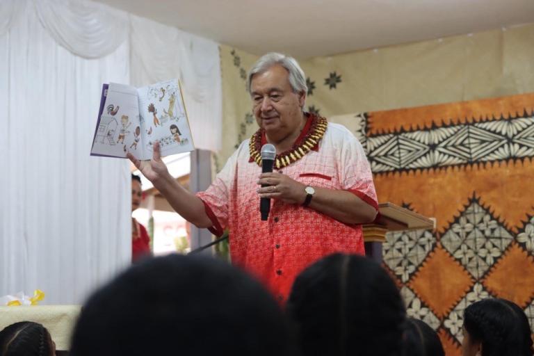 A man wearing a red and white shirt holds an open book while speaking to an audience. The room has patterned decorations on the walls.