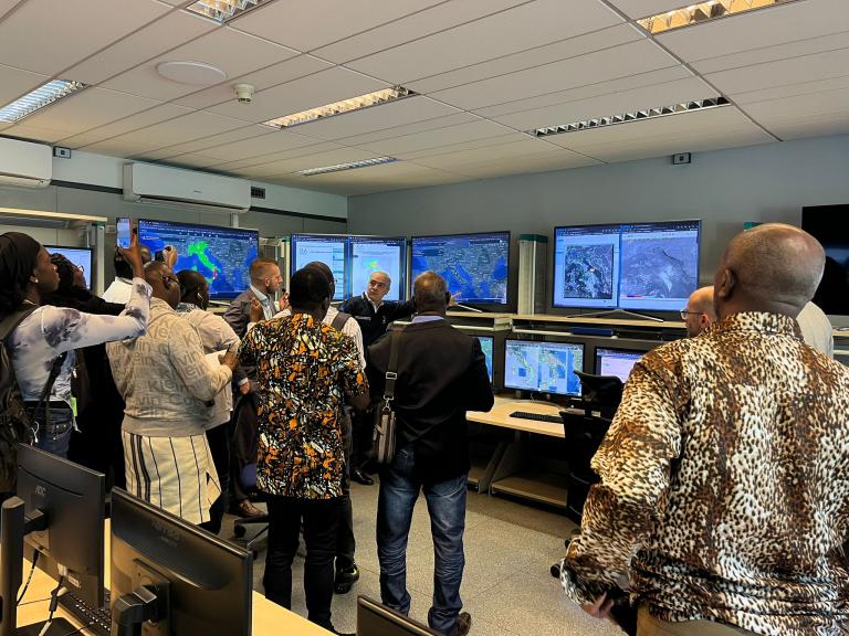 A group of people in a control room observes several large monitors displaying maps and data.