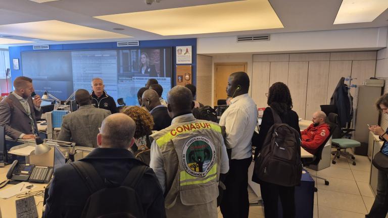 A group of people stand in a room, some wearing vests with logos. A screen and various office equipment are visible in the background.