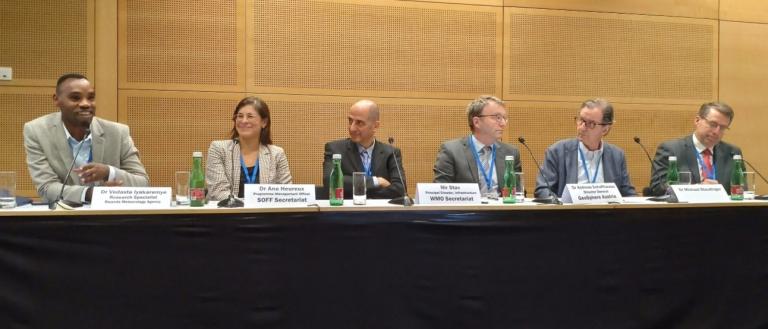 A panel of six people seated behind a table, each with microphones and name placards. Bottles of water are also on the table.