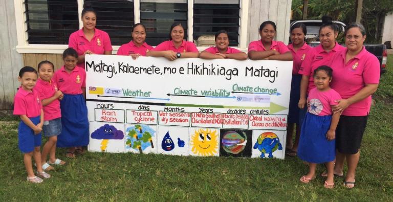 A group of women and children in pink shirts stand around a sign about weather, climate variability, and climate change, with drawings and text detailing various environmental factors.