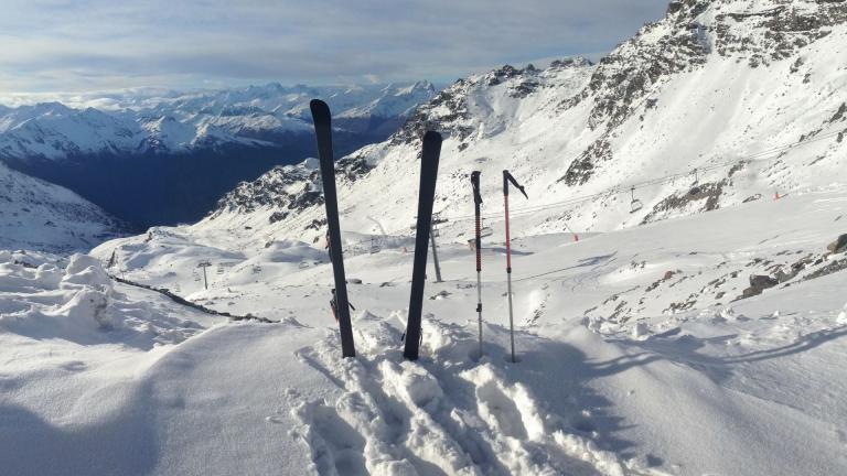 A snowy mountain landscape with two pairs of skis and poles stuck in the snow, overlooking a range of distant peaks under a partly cloudy sky.