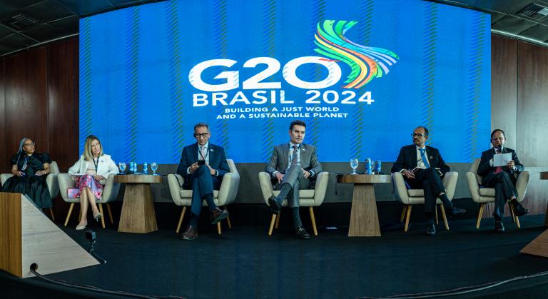 Panel discussion at G20 Brasil 2024 event, featuring six individuals seated on stage with a large screen displaying the event logo behind them.