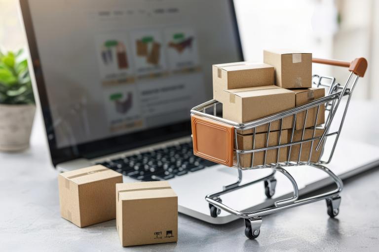 Small cardboard boxes in a miniature shopping cart in front of a laptop showing an online shopping page.