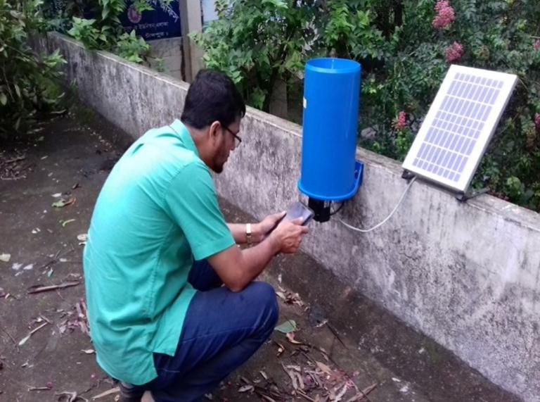 A person in a green shirt checks a blue rain gauge attached to a wall with a small solar panel nearby.