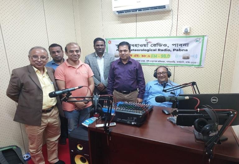 A group of six men standing in a radio studio with broadcasting equipment and a banner on the wall.