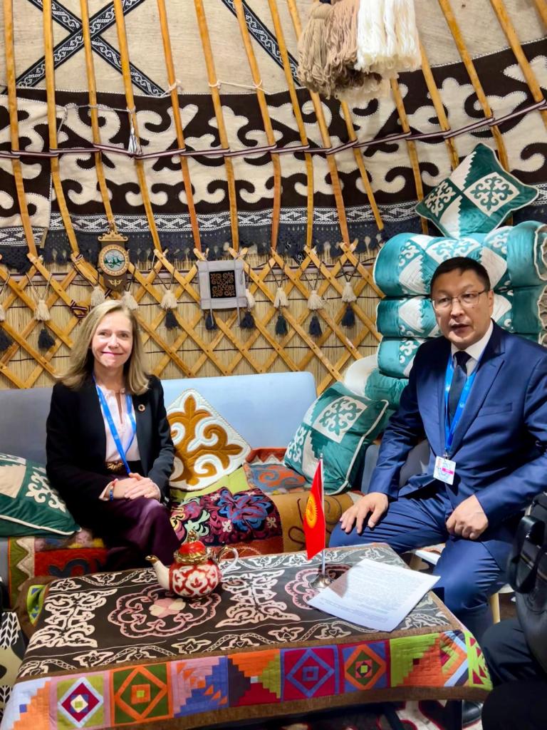 Two people sit inside a decorated yurt, adorned with colorful textiles and cushions. A small Kyrgyz flag is on the table in front of them.