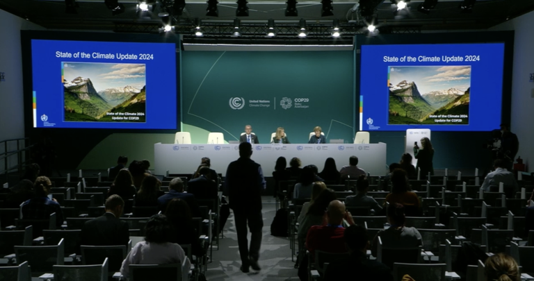 A conference room with three speakers on stage discussing "State of the Climate Update 2024." The audience is seated, and the presentation screen displays a mountain landscape.