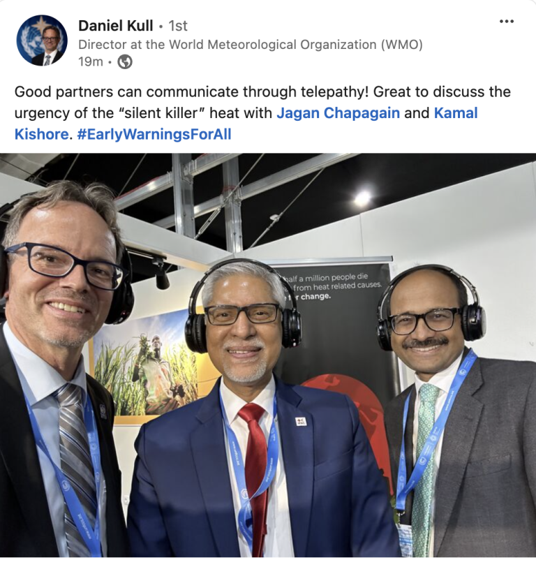 Three men wearing headphones stand together, smiling, at a World Meteorological Organization event. A poster about heat-related deaths is visible in the background.