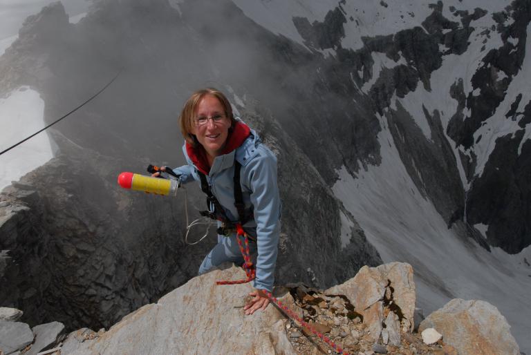 A climber in a blue outfit stands on a rocky peak, holding an avalanche probe pole, with snow and clouds in the background.