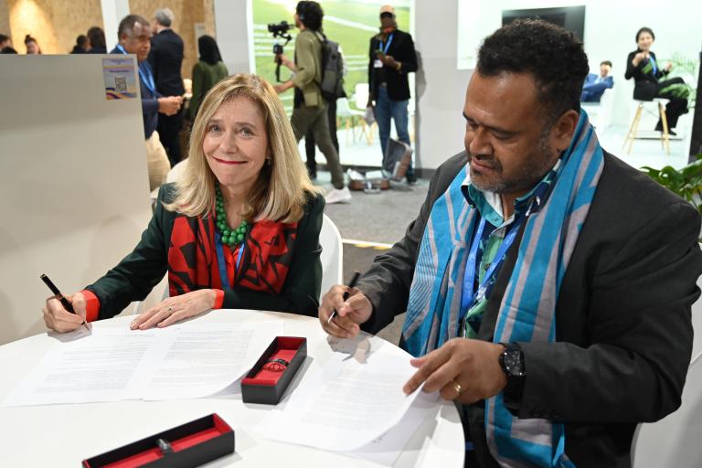 Two people sitting at a table signing documents, with pens in hand. The woman wears a green jacket, and the man wears a blue scarf. A group of people is visible in the background.