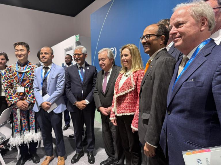 A group of people in formal attire pose for a photo in front of a blue wall. Some are wearing suits, while one person wears a colorful, traditional outfit.