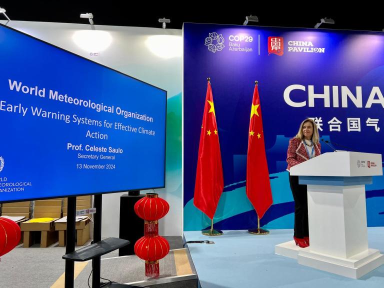 A speaker stands at a podium next to a screen displaying a presentation on early warning systems for climate action at COP29, with the Chinese national flags in the background.