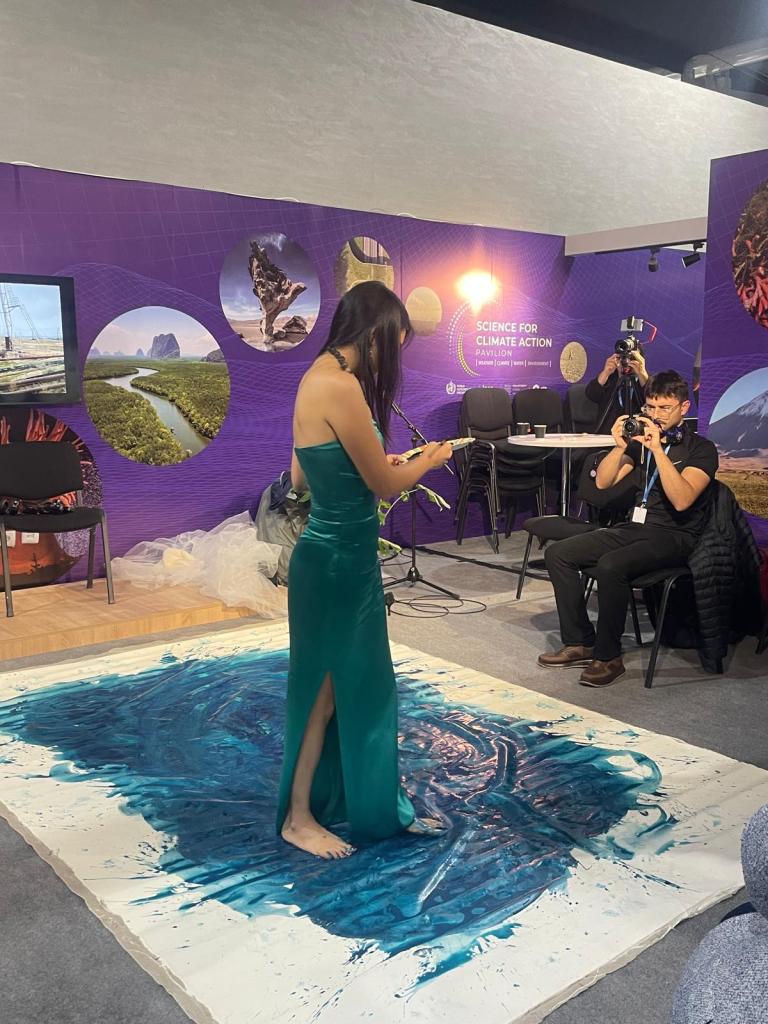 A woman in a green dress stands barefoot on a canvas with blue paint, surrounded by photographers at an event.