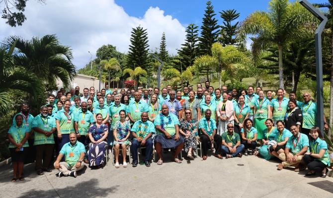 A large group of people wearing green shirts pose outdoors in front of palm trees on a sunny day.