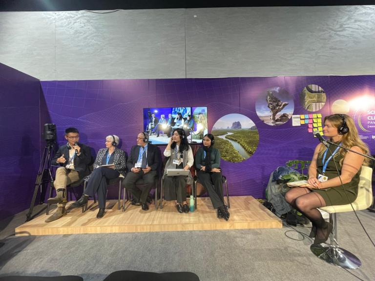 Panel discussion with six people seated on a stage, microphones in use, and a woman taking notes. A screen displays images behind them.