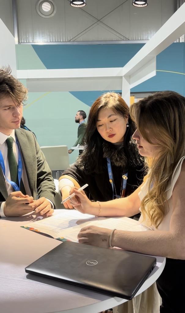 Three people are discussing at a table with documents and a laptop.