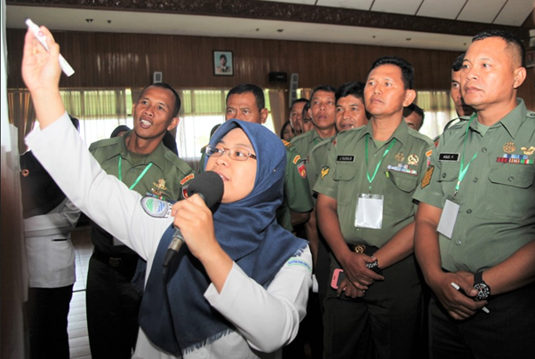 A woman in a hijab holds a microphone and points at a board, addressing a group of uniformed military personnel in a dimly lit room.