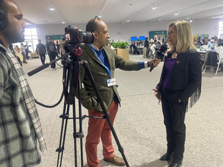 A woman is being interviewed by a reporter in a conference room. A cameraman is recording the interview. People are seated at tables in the background.