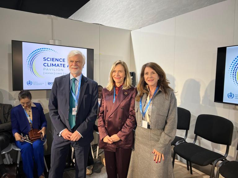 Three people stand in front of a "Science & Climate Pavilion" sign. Two are women and one is a man, all wearing business attire. Three chairs and two screens are visible in the background.