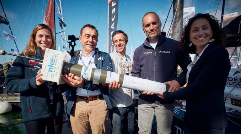 Five people stand together, smiling and holding a large rolled document or tube, with boats and flags visible in the background.