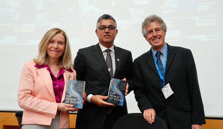 Three people in business attire holding a book titled "The Primacy of Doubt," standing in front of a presentation screen.