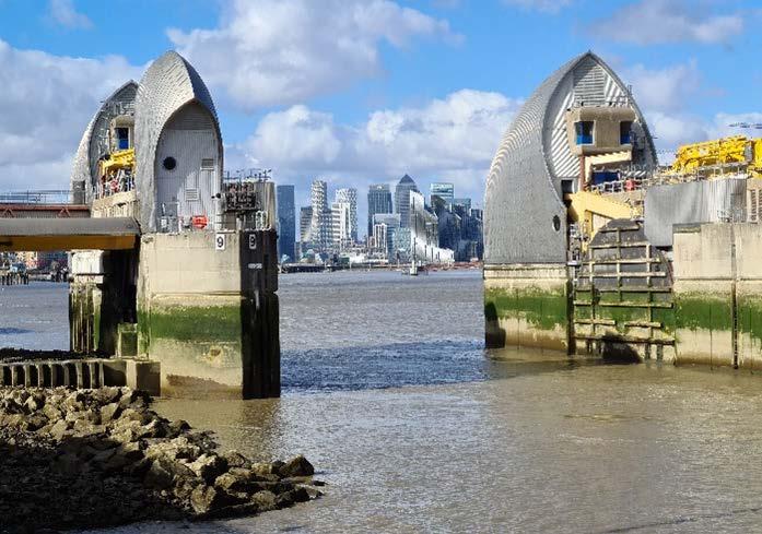 The image shows the Thames Barrier, consisting of large silver gates partially open on a river, with a city skyline in the background under a cloudy sky.