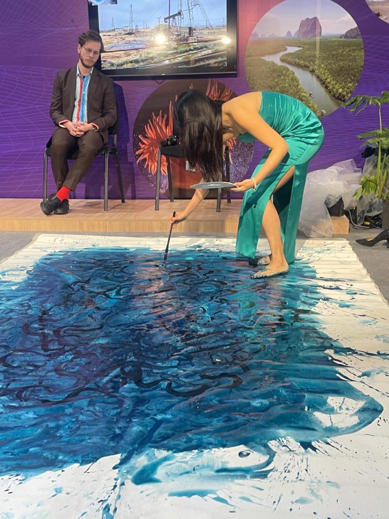 A woman in a green dress paints on a large canvas on the floor while a man in a suit observes. A screen and plant images are in the background.