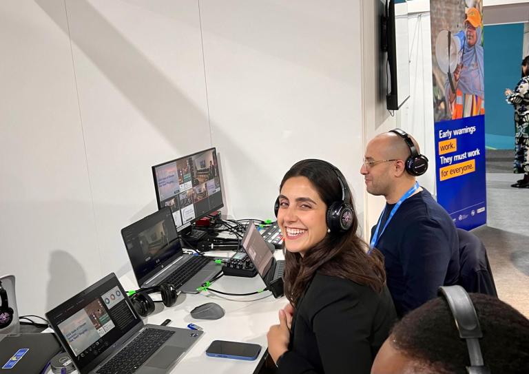 People wearing headsets work at a control desk with monitors. A poster in the background reads, "Early warnings work. They must work for everyone.